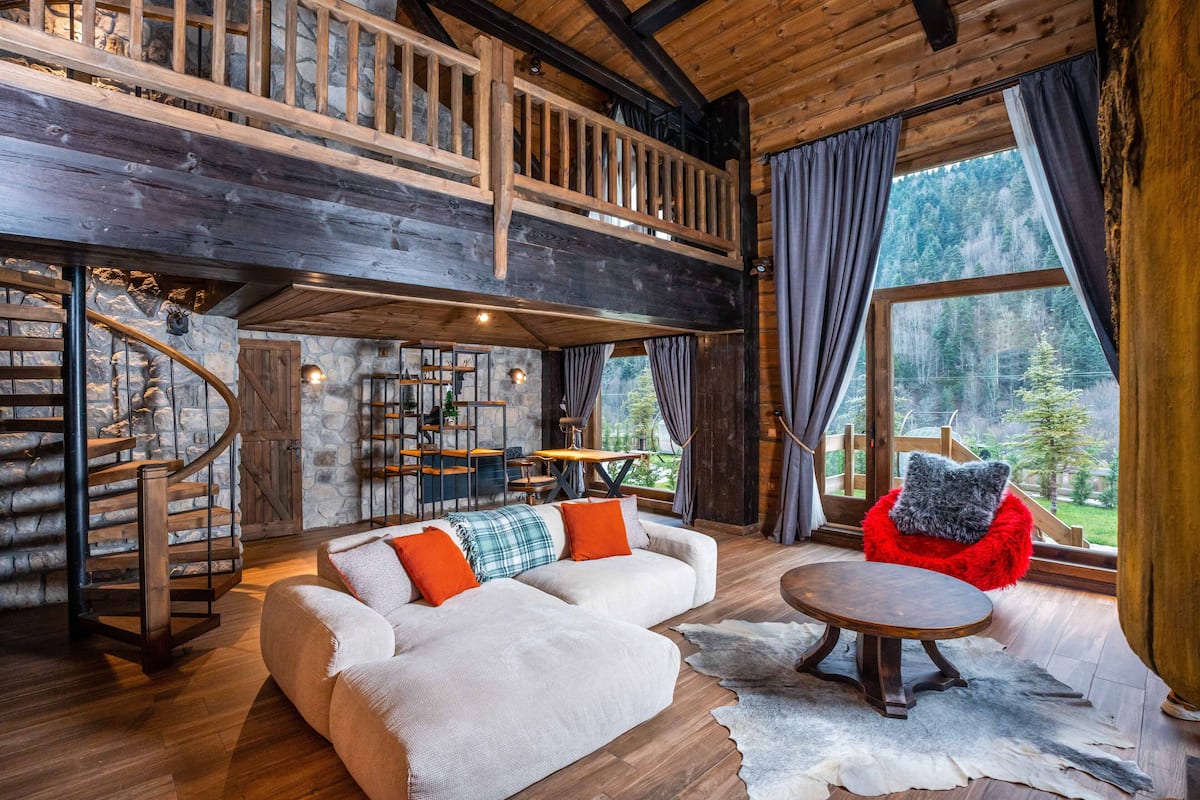 A spacious living area with a wooden staircase, a sofa with orange cushions, a red chair, and a large window offering a forest view.