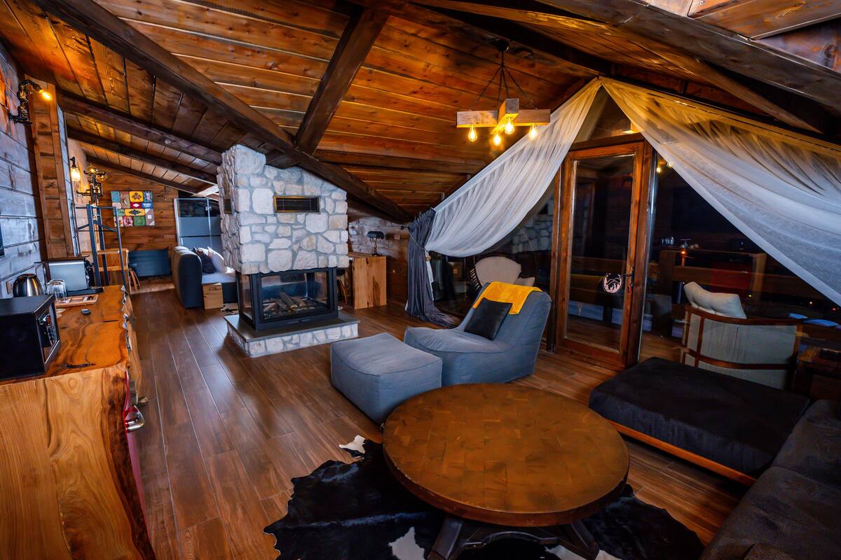 A cozy loft-style room with a stone fireplace, wooden beams, and a comfortable seating area with a sofa and armchairs.