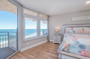 A bedroom with a large window overlooking the ocean, a bed with a nautical-themed bedspread, and a sign that reads "PARADISE" on the wall. - Blue Surf 705 - Daytona Beach Oceanfront Vacations, Inc (Daytona Beach Shores)