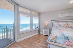 A bedroom with a large window overlooking the ocean, a bed with a nautical-themed bedspread, and a sign that reads "PARADISE" on the wall.
