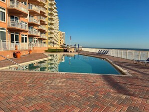 Une piscine avec une terrasse en briques, entourée d’une clôture, et un bâtiment avec des balcons en arrière-plan.
