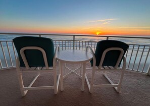 Una mesa y dos sillas en un balcón con vista al atardecer sobre el agua.