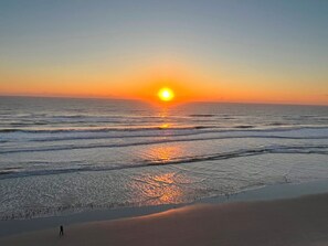 Une plage où le soleil se couche sur l’océan, les vagues venant doucement lécher le rivage, et une personne qui se promène le long de la plage.