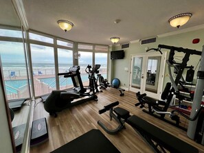 A gym with cardio machines, weightlifting equipment, and a bench. It has large windows with a view of the ocean and a pool. - Blue Surf 705 - Daytona Beach Oceanfront Vacations, Inc (Daytona Beach Shores)