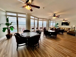 A spacious living room with a large dining table, multiple chairs, a sofa, and a television. There are large windows offering a view of the ocean. - Blue Surf 705 - Daytona Beach Oceanfront Vacations, Inc (Daytona Beach Shores)
