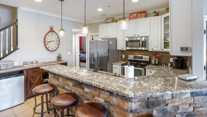 Une cuisine moderne dotée de plans de travail en granit, d’un îlot central en pierre, de tabourets de bar, d’appareils électroménagers en acier inoxydable et de luminaires suspendus.