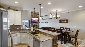 Une cuisine moderne dotée de plans de travail en granit, d’un îlot central intégré, d’un mur d’accent en pierre et d’un coin repas avec des chaises et une table en bois.