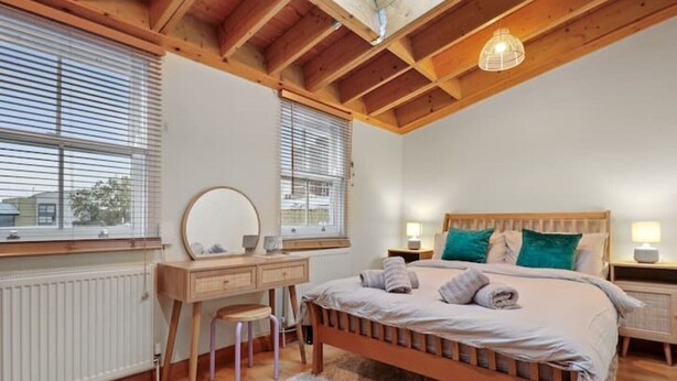 Een slaapkamer met een houten bed, een houten dressoir, een spiegel en een raam met jaloezieën.