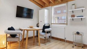Een moderne, goed verlichte kamer met een houten plafond, een ronde tafel, twee stoelen, een televisie, een radiator en een raam met zonwering.