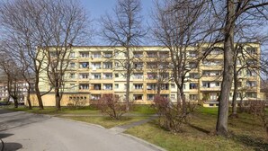 A multi-story residential building with balconies, surrounded by bare trees and a paved area in front.