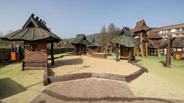 A rustic wooden play area with multiple small structures, including a central building and several smaller huts with conical roofs.