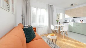 A modern living room with an orange sofa, a dining area with a white table and chairs, and a kitchen with light green cabinets.