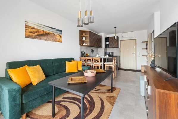 Un salon moderne avec un canapé vert, des coussins jaunes, une table basse et un téléviseur posé sur un meuble en bois.