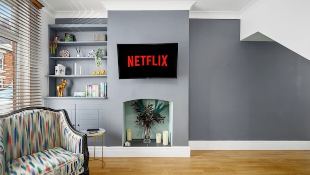 Una sala de estar moderna con una pared gris, un televisor montado mostrando 'NETFLIX', una estantería, un sofá estampado y una chimenea.