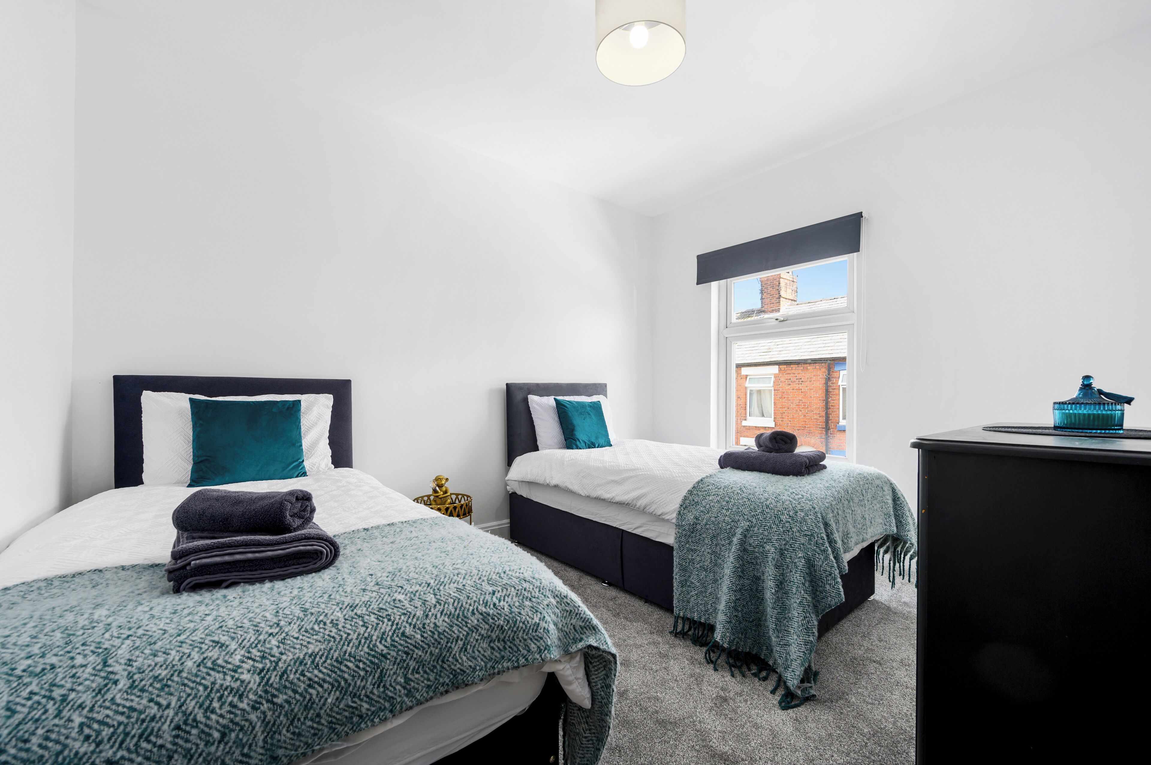 A bedroom with two beds, each with a dark headboard, white bedding, and teal throw pillows. There is a window with a view of a brick building, a desk with a lamp, and a small decorative item on the desk.