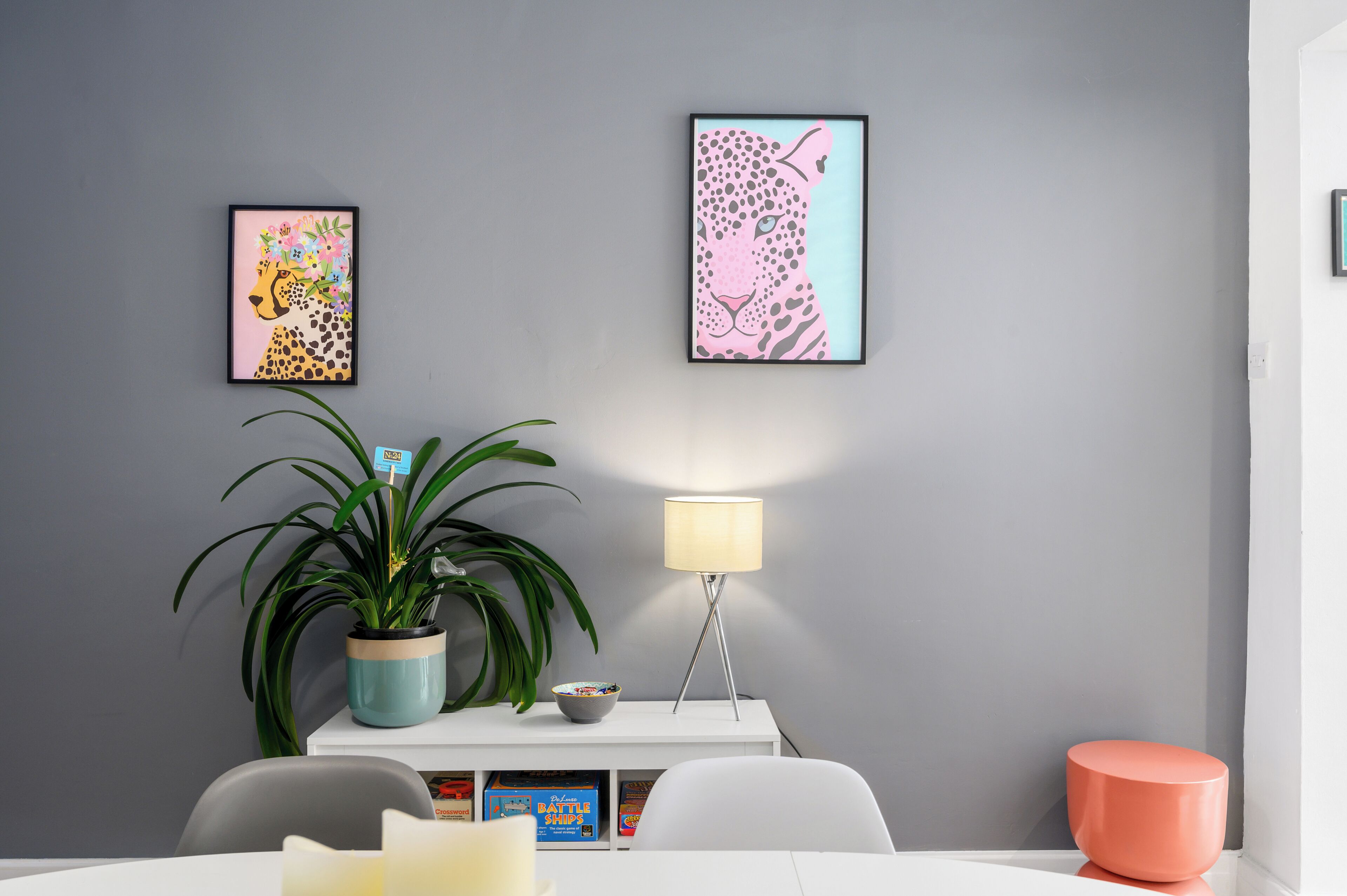 Una habitación moderna con pared gris, dos cuadros enmarcados, una planta en maceta, una lámpara de pie, una mesita con un cuenco y una silla blanca.