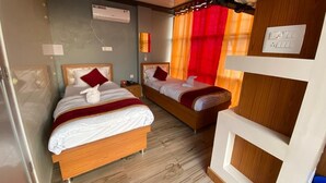 A hotel room with two wooden beds, each with white bedding and red pillows. There is a ceiling fan, an air conditioning unit, and a light switch on the wall.