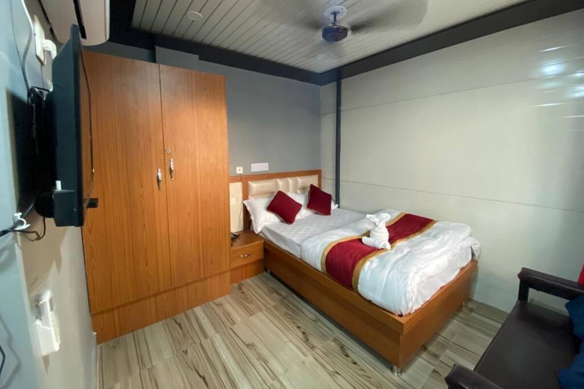 A bedroom with a wooden wardrobe, a bed with white bedding and red accents, a ceiling fan, and a wooden floor.