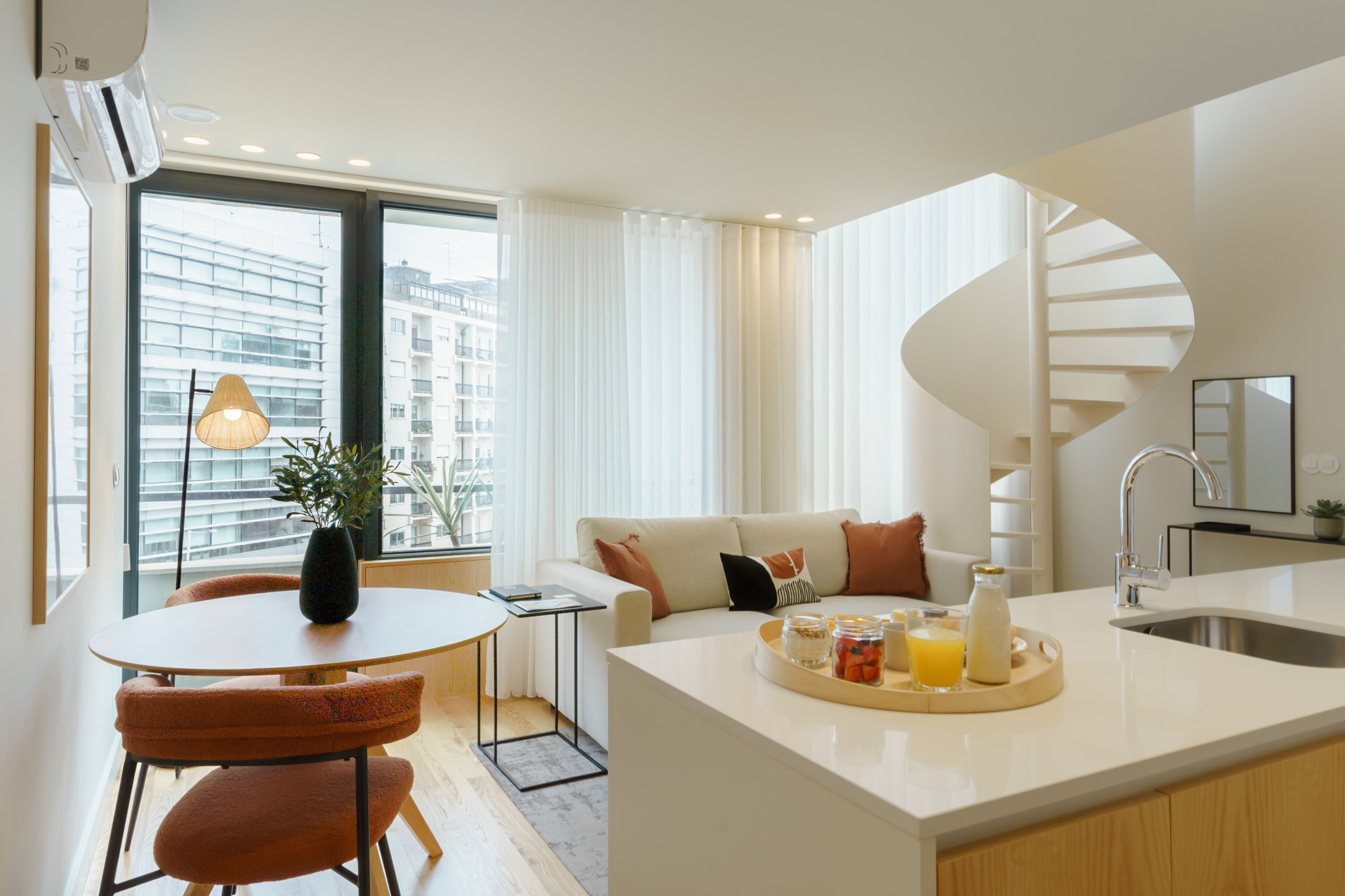 A modern kitchen with a central island, a dining area with orange chairs, a spiral staircase, and a view of buildings through large windows.