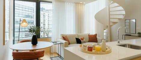 A modern kitchen with a central island, a dining area with orange chairs, a spiral staircase, and a view of buildings through large windows.