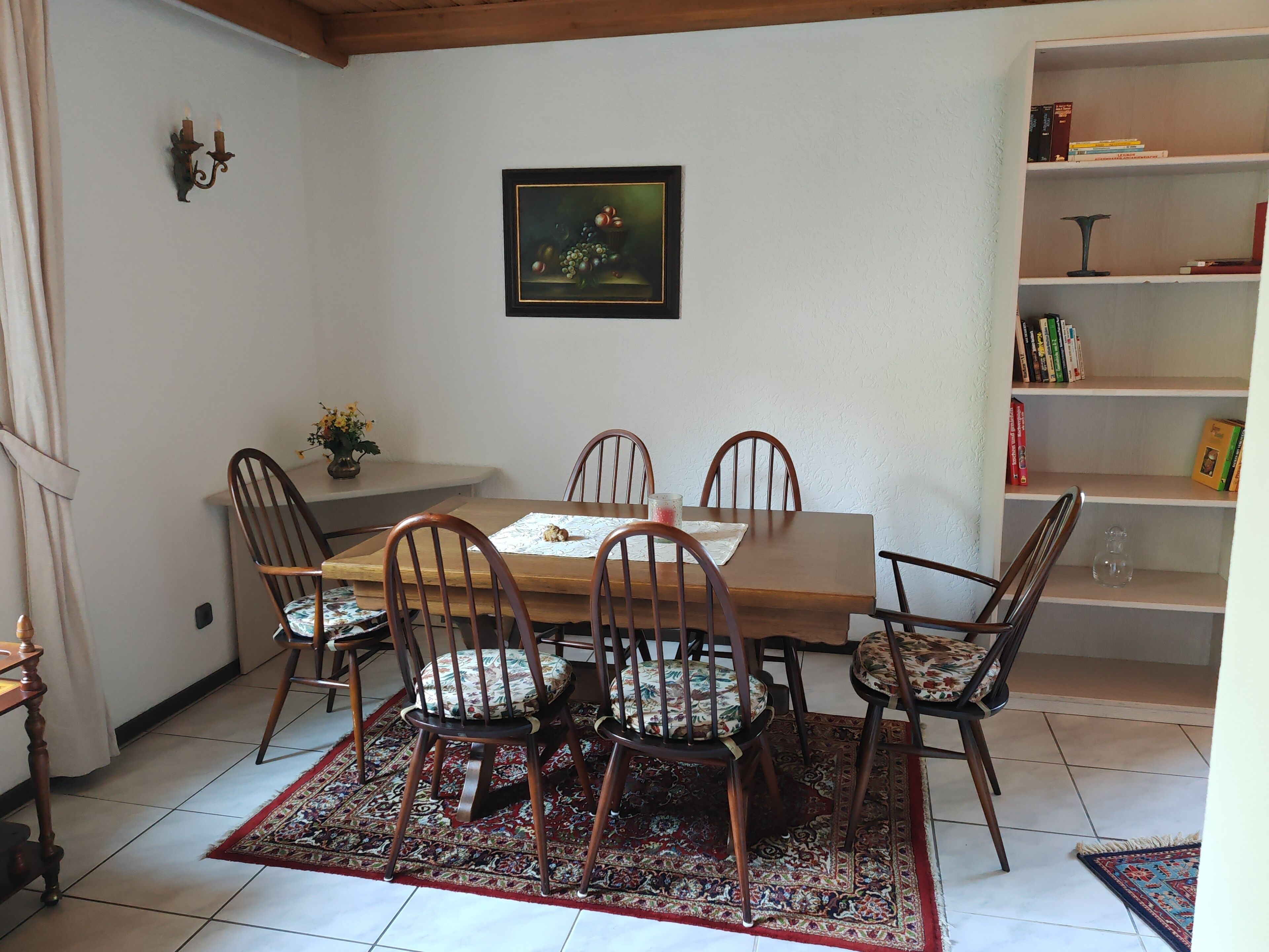 Un coin repas avec une table et des chaises en bois, un tapis à motifs, un tableau au mur et une bibliothèque contenant des livres et des objets décoratifs.