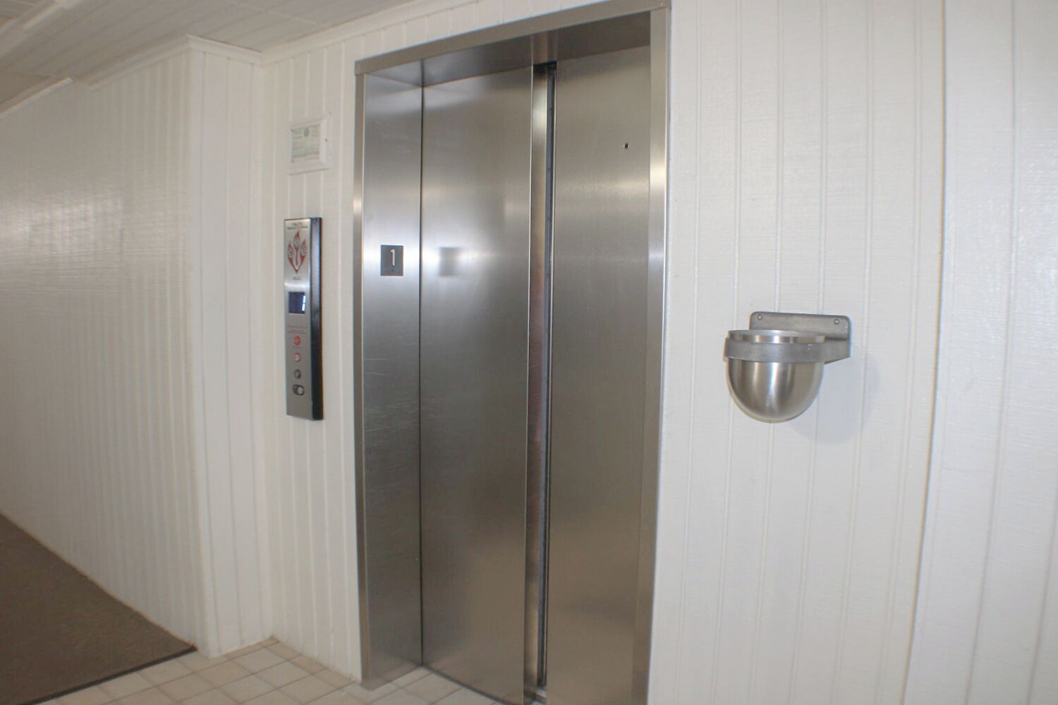 A metallic elevator with a control panel and a wall-mounted light fixture in a hallway with white paneling.