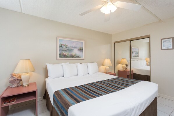 A hotel room with a bed, two bedside tables, a ceiling fan, a painting on the wall, and a mirror.