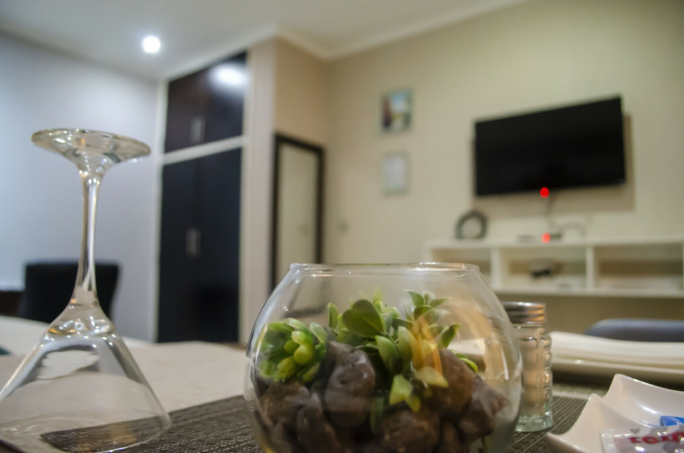 A living room with a glass table, a glass vase with plants, a wine glass, and a flat-screen TV mounted on the wall.