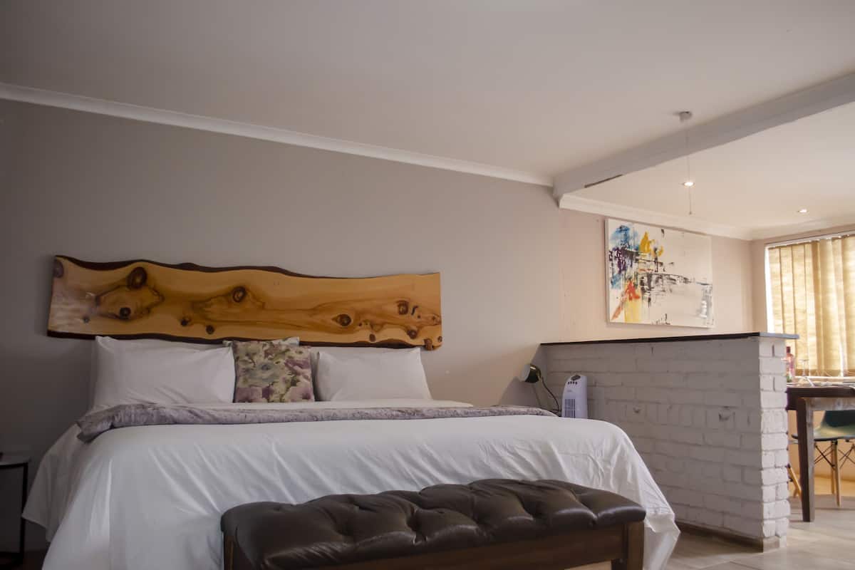 Quarto com uma cama grande, cabeceira de madeira, parede branca, um quadro e uma janela com cortinas.