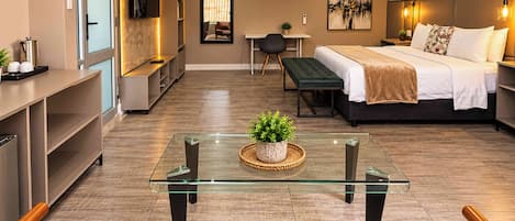 A modern hotel room with a glass coffee table, a bed with a green upholstered bench, a desk with a chair, and a TV mounted on the wall.