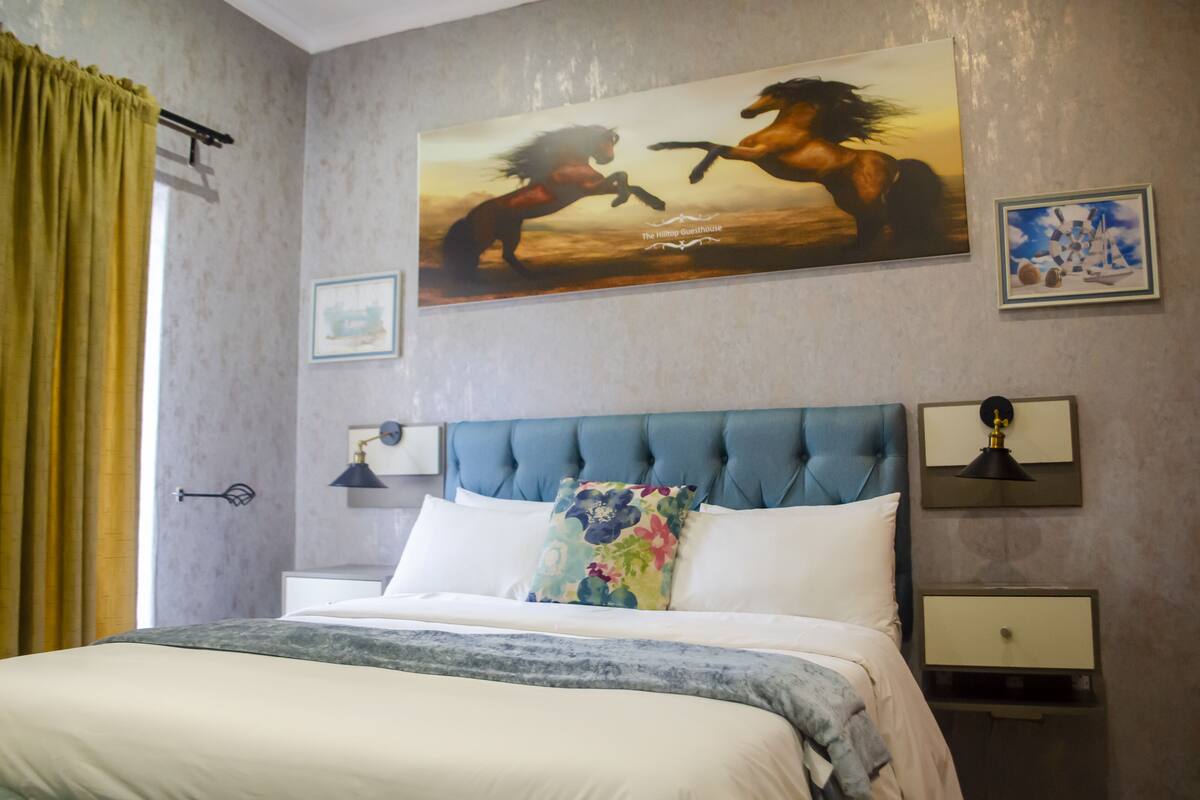 Quarto de hotel com uma cama grande, cabeceira azul, duas mesas de cabeceira com luminárias, janela com cortinas e um quadro com cavalos na parede.