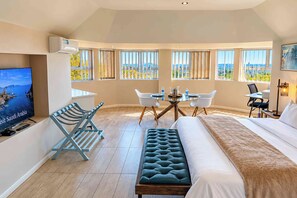 Habitación de hotel con cama, escritorio, silla, banco, televisor y ventanas grandes con vistas.