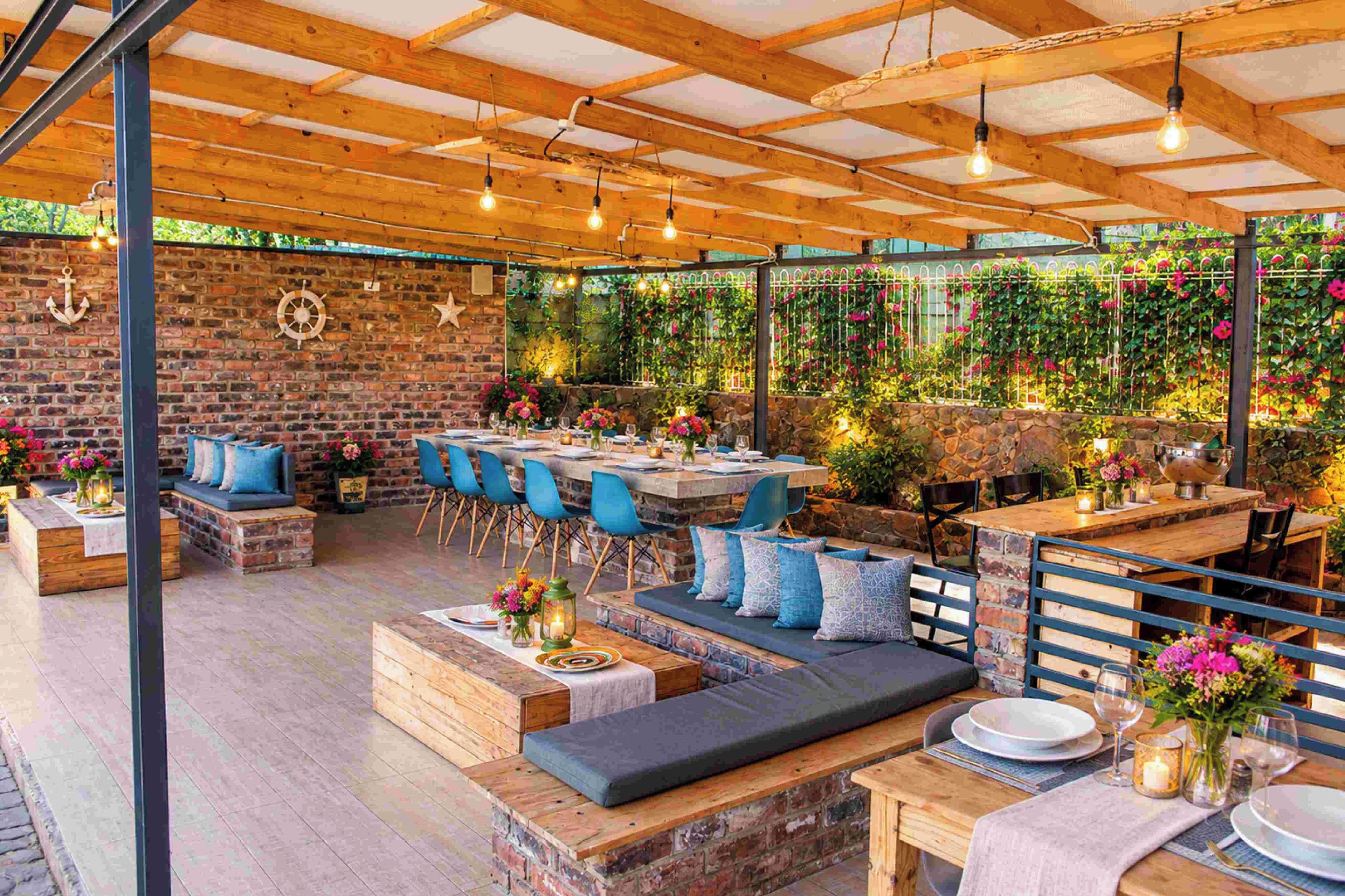 An outdoor patio with wooden tables and blue chairs, stone walls, and hanging plants.