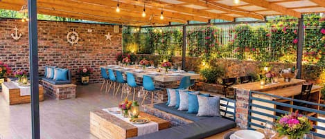Un patio al aire libre con mesas de madera, sillas azules, muros de piedra y plantas colgantes.