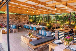 An outdoor patio with wooden tables and blue chairs, stone walls, and hanging plants.
