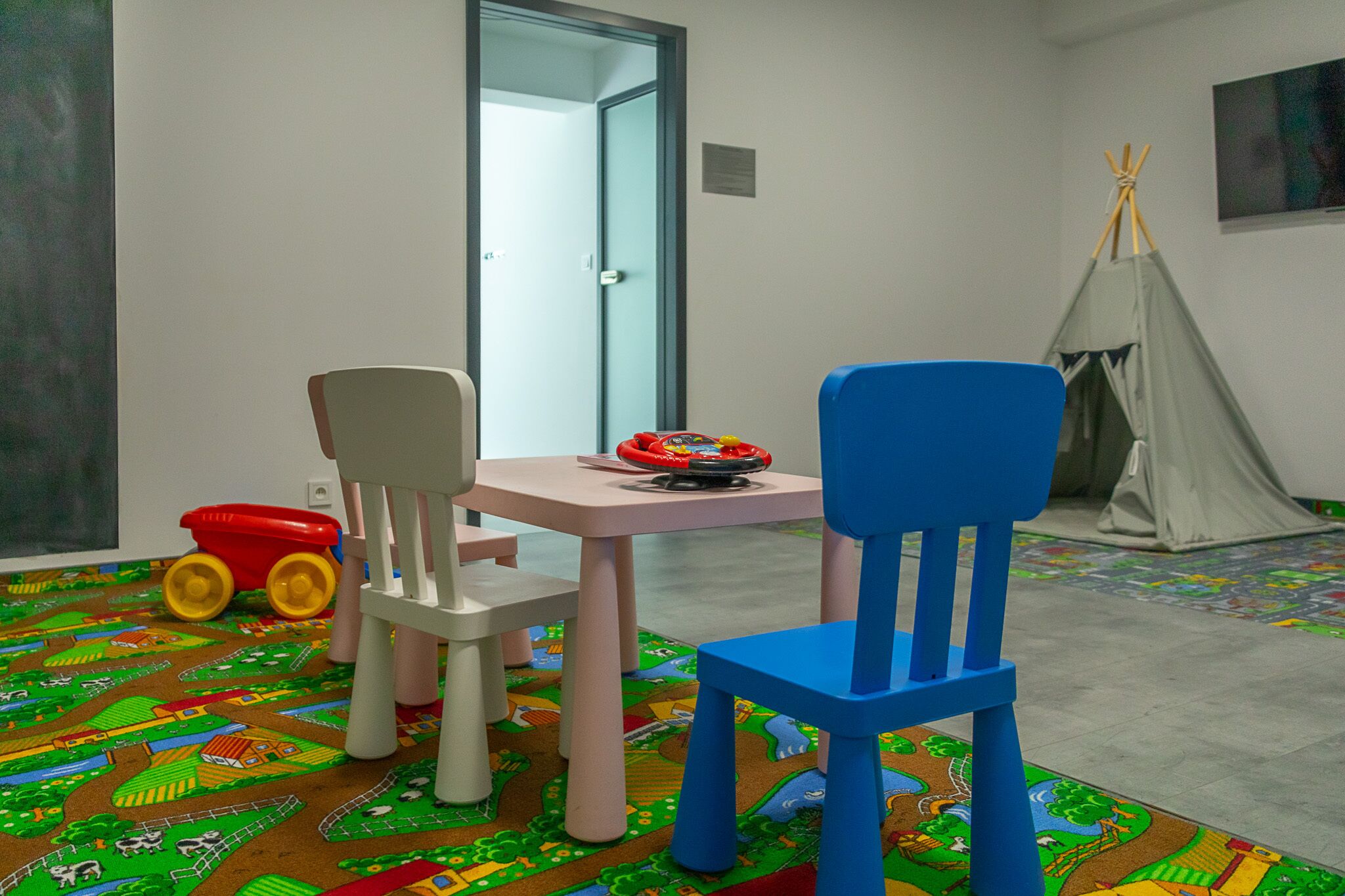 Um quarto com área de recreação, incluindo um carrinho de brinquedo vermelho, duas cadeiras, uma mesa e um tipi.