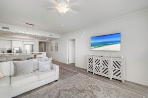 A modern living room with a white sofa, a patterned rug, a ceiling fan, a kitchen area, and a large beach-themed painting on the wall. - Perdido Dunes Tower 1W by Liquid Life (Orange Beach)