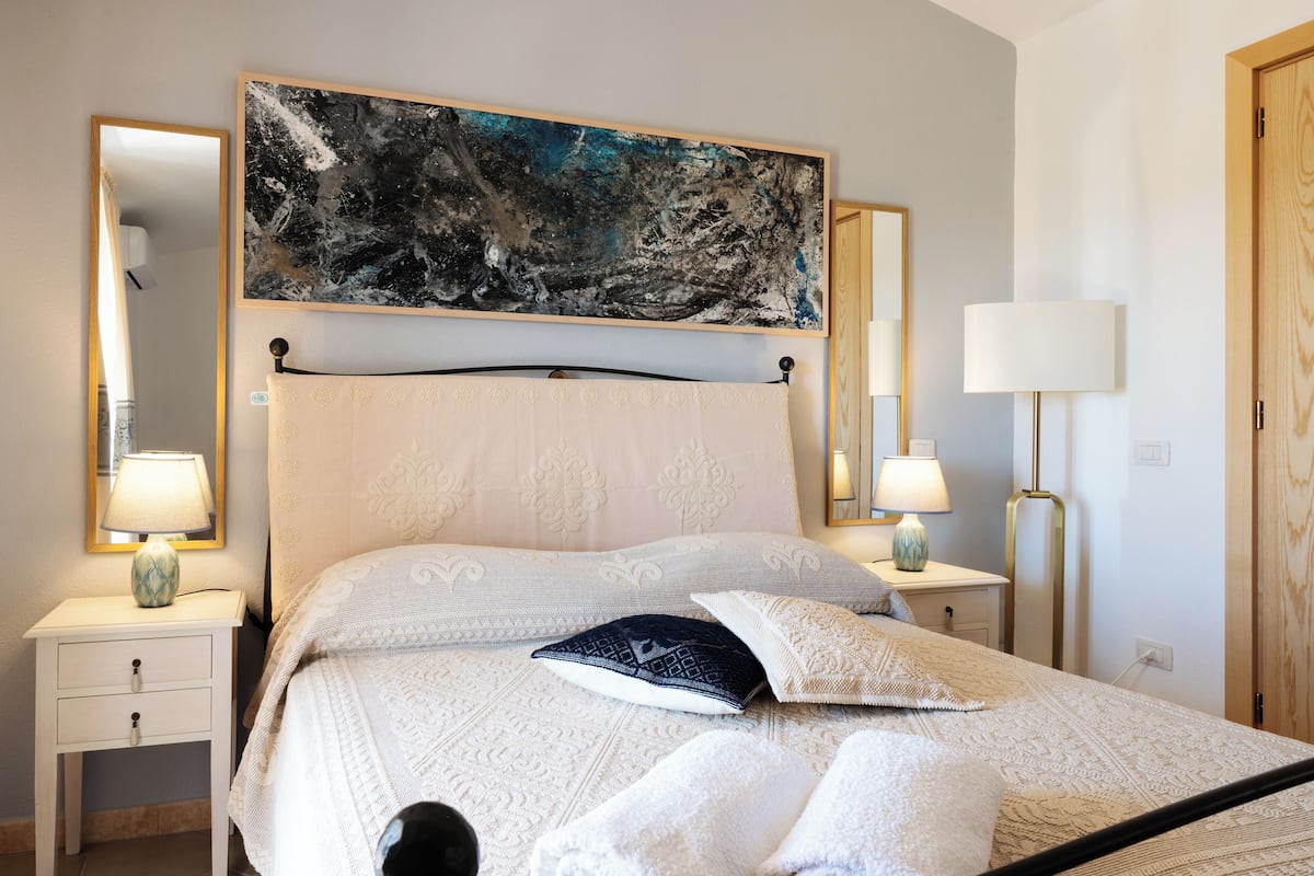 Una cama bien hecha con una colcha estampada, dos lámparas de noche y un cuadro abstracto grande sobre el cabecero.