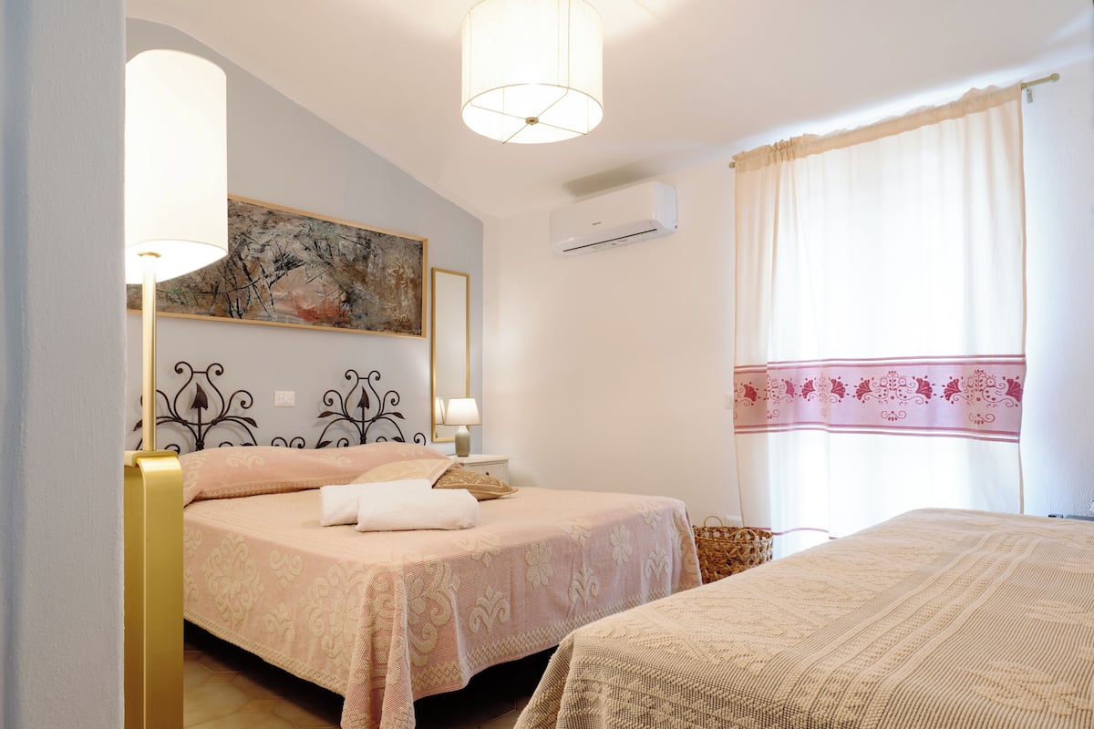 Un dormitorio con dos camas, una mesita de noche, una lámpara, aire acondicionado y una ventana con cortinas.