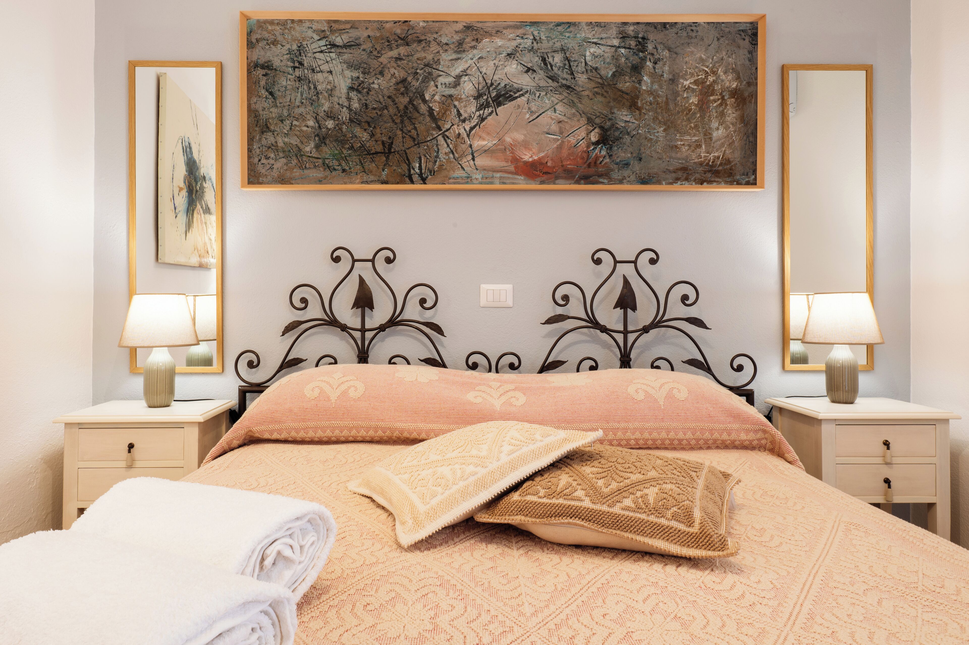 A neatly made bed with patterned bedding, two bedside tables with lamps, and a large abstract painting above the bed.