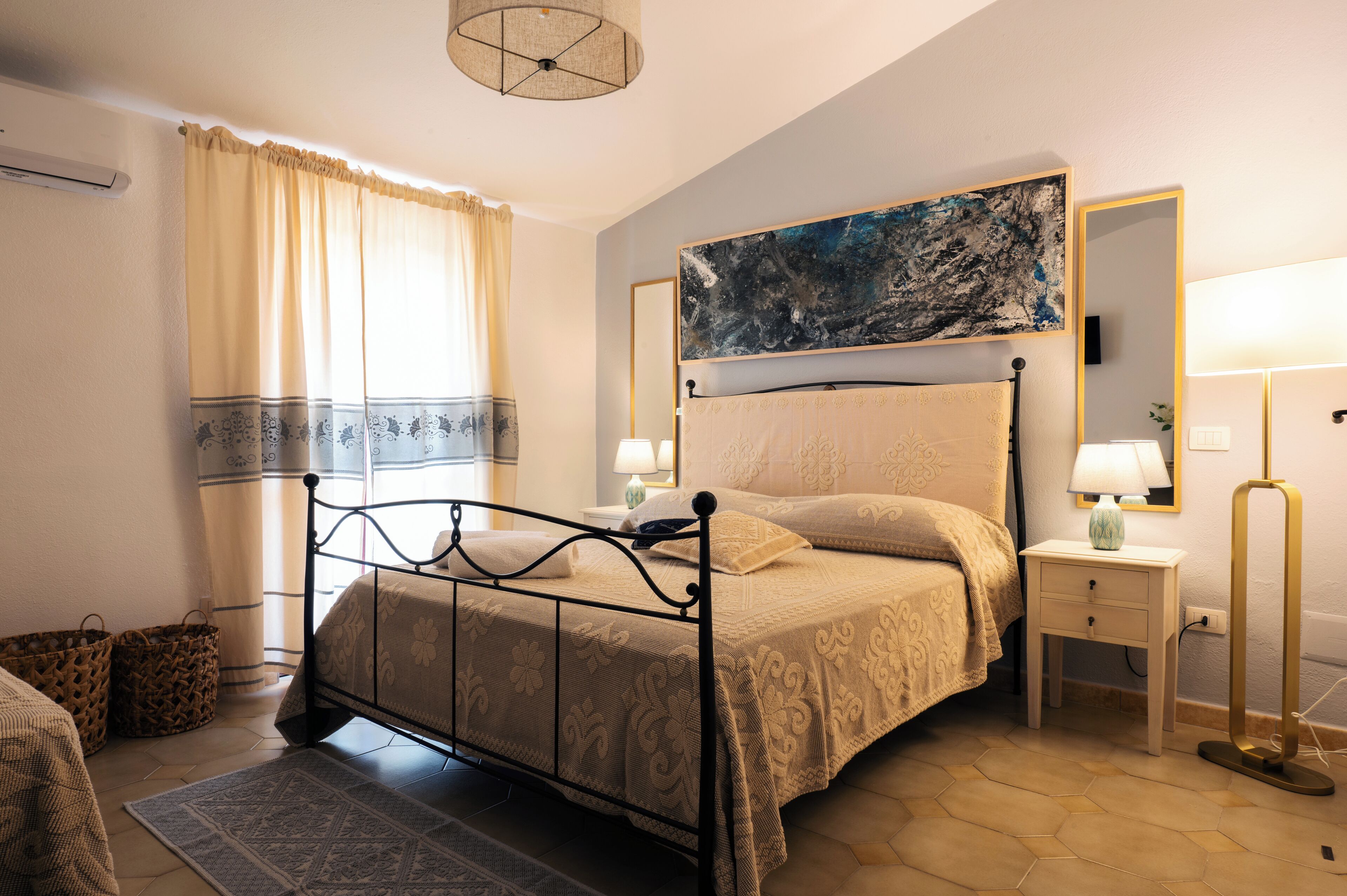 Un dormitorio con cama, mesitas de noche, una lámpara, un cuadro y una ventana con cortinas.