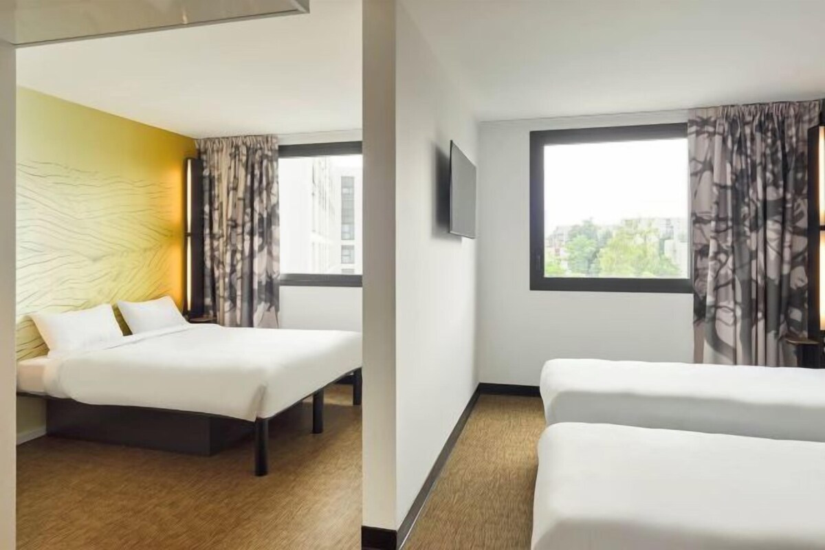 A hotel room with a bed, a window with curtains, a flat-screen TV, and a view of buildings outside.