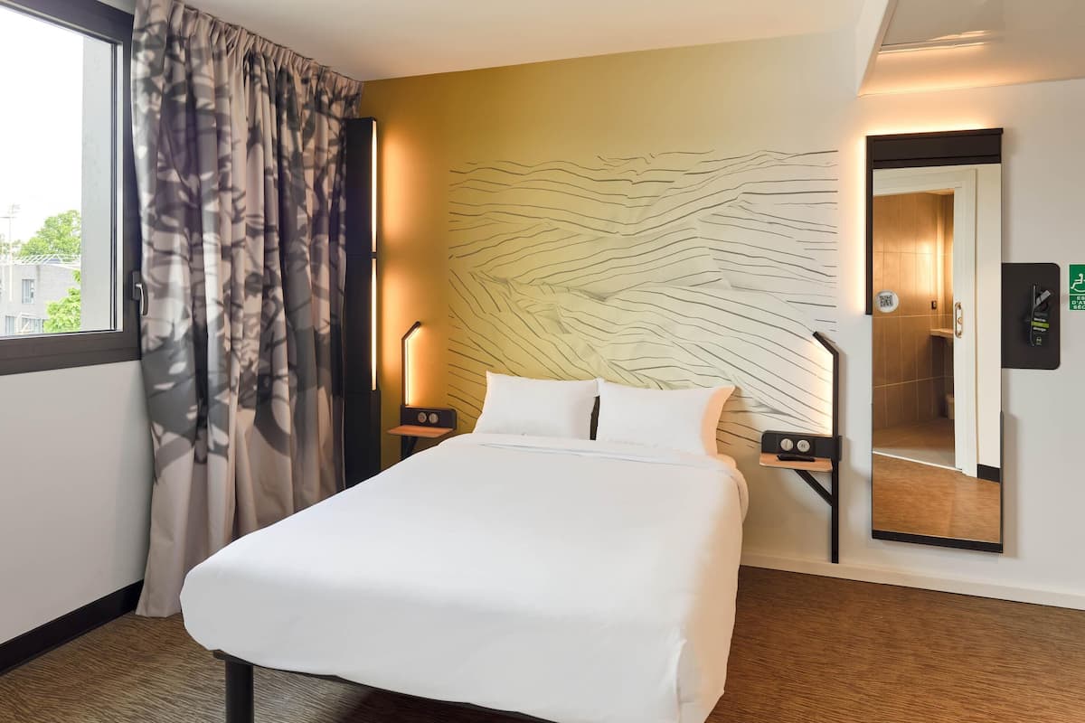 Ein Hotelzimmer mit einem großen Bett, einem Wandspiegel, einem Nachttisch mit Lampe und einem mit Vorhängen drapierten Fenster.