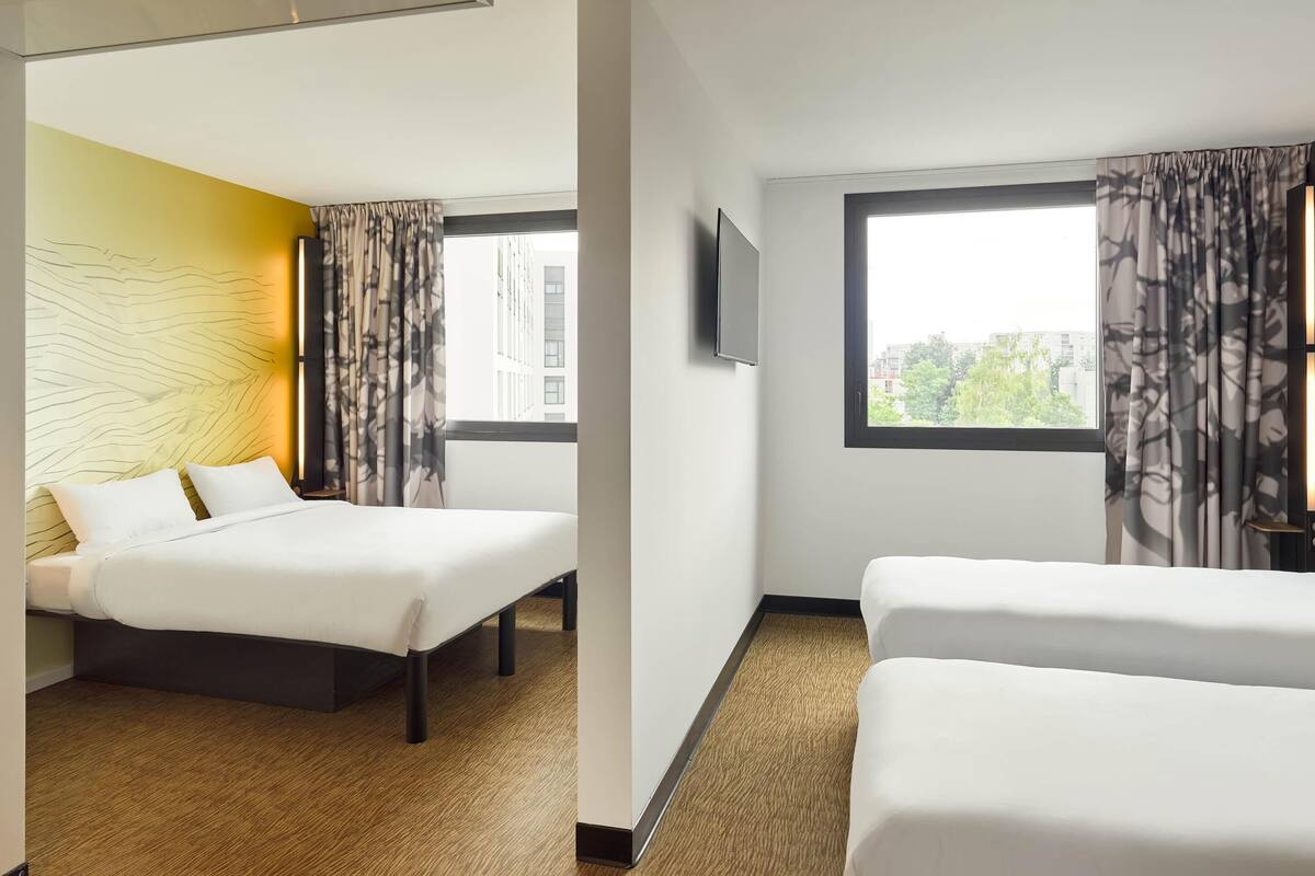 Ein Hotelzimmer mit zwei Betten, einem großen Fenster mit Vorhängen, einem Flachbildfernseher und einer gelben Akzentwand.