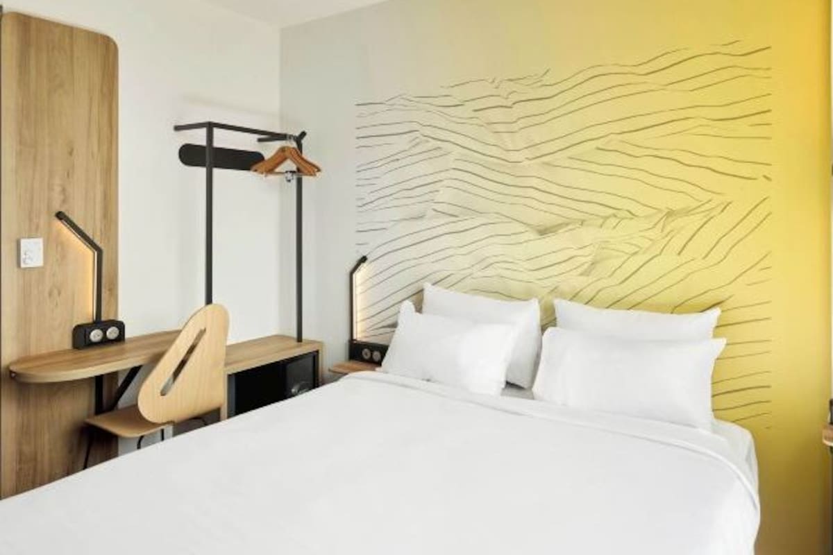 Ein Hotelzimmer mit Bett, Schreibtisch, Stuhl, Kleiderständer und einer Wand mit strukturierter Gestaltung.