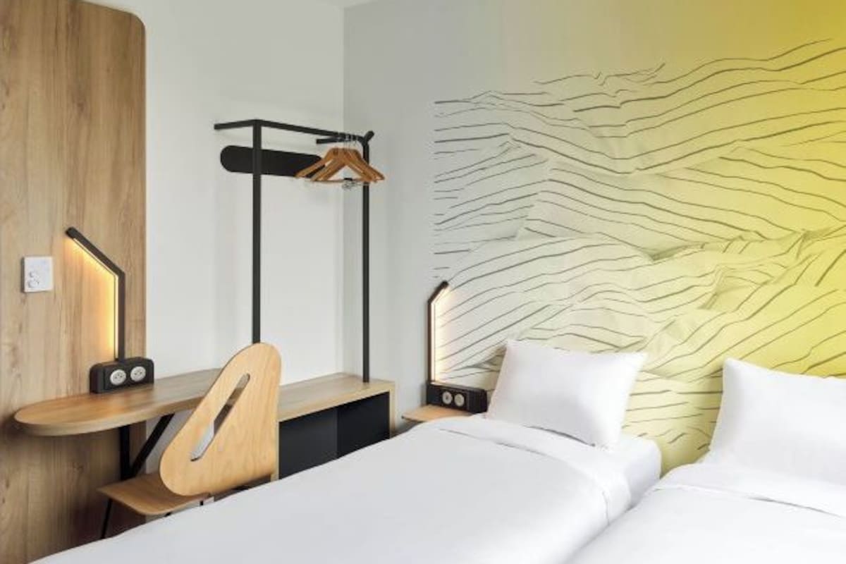 Ett modernt hotellrum med ett skrivbord i trä, en klädstång, en säng med vita sängkläder och en vägg med ett vågigt mönster.