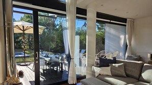 A modern living room with a large glass door leading to an outdoor patio. The room features a grey sofa, a small ottoman, and a coffee table. There is a dining area visible through the glass door with outdoor furniture and a pergola. - Troia Beach House With Pool (Grandola)