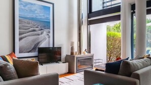 A modern living room with a large TV, comfortable seating, a bookshelf, and a view of greenery through large windows. - Troia Beach House With Pool (Grandola)