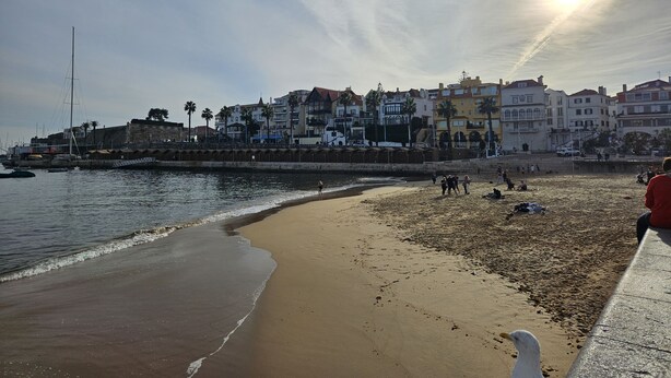 A coastal beach with a stone wall, a sailboat, and buildings in the background. - Historic Center Cascais Bolina 2 (Cascais)