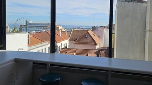 Ett rum med ett stort fönster som vetter ut mot en stadsbild, med utsikt över byggnader och ett pariserhjul. I förgrunden står två pallar.